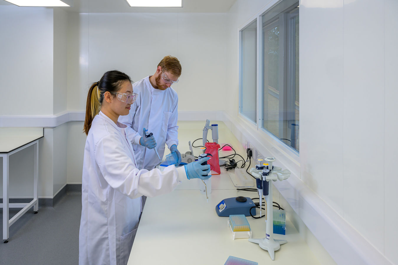 Two scientists in lab coats and safety goggles use pipettes at a lab bench, surrounded by various laboratory equipment and supplies, in a bright, sterile laboratory environment.