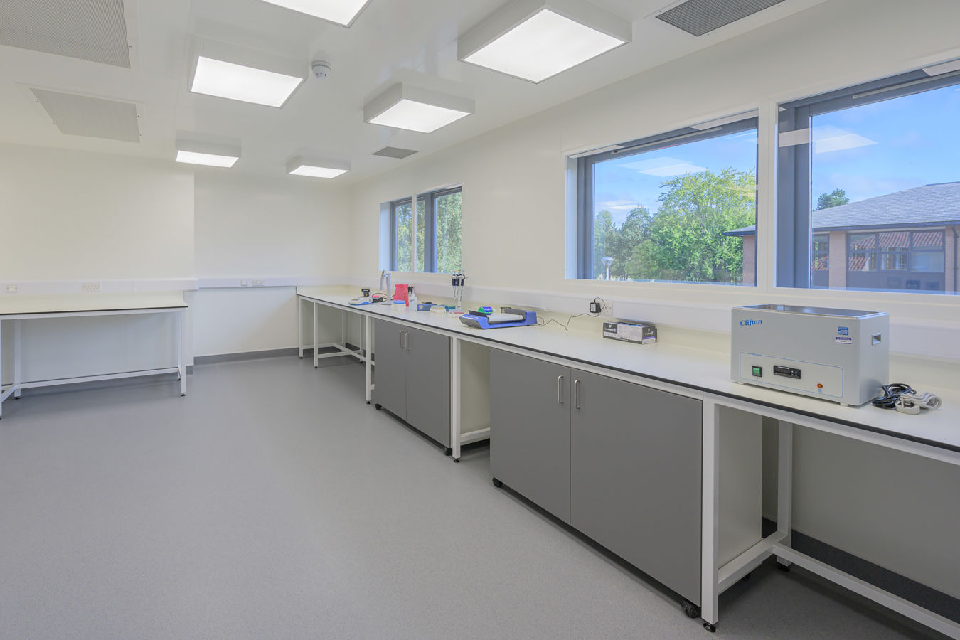 Bright laboratory with grey cabinets, white countertops, and large windows providing natural light and views of greenery outside.