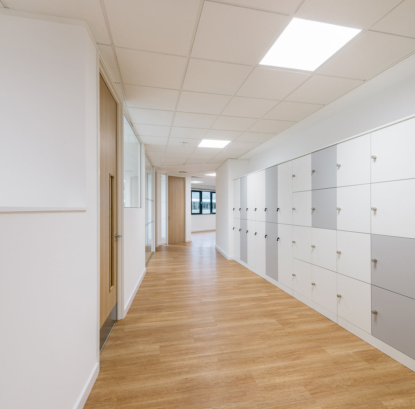 Clean and modern hallway with wooden flooring, white lockers along one side, and ceiling lights creating a bright, organized space.