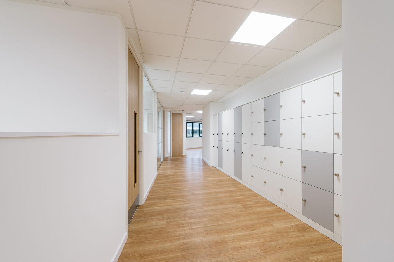 Clean and modern hallway with wooden flooring, white lockers along one side, and ceiling lights creating a bright, organized space.