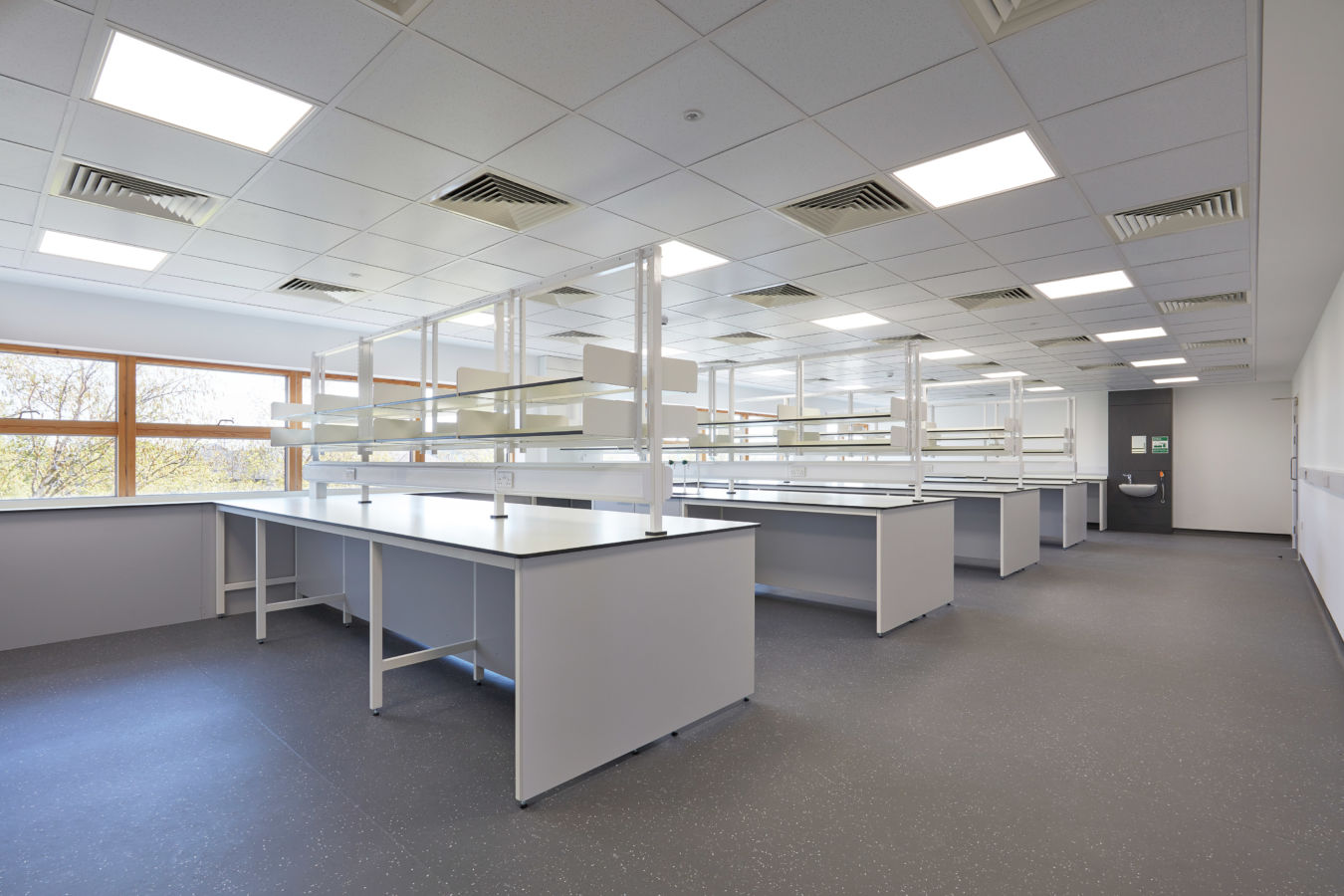 Spacious laboratory with multiple white workstations, overhead shelves, and a grid ceiling with recessed lighting and ventilation panels.
