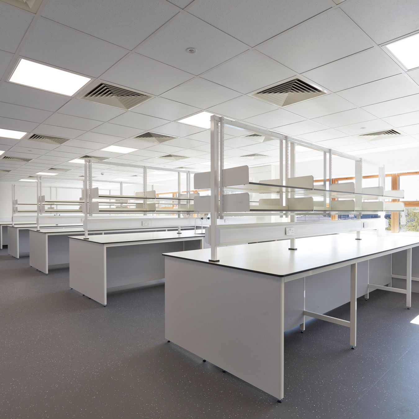 Spacious, well-lit laboratory with multiple white workbenches, overhead shelving units, and a grid of fluorescent ceiling lights.