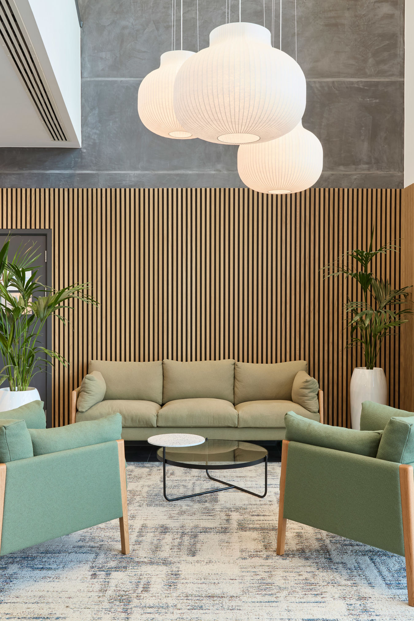 A modern lobby features green armchairs and a sofa around a glass coffee table, with large plants and pendant lights, surrounded by wooden paneling and large windows.