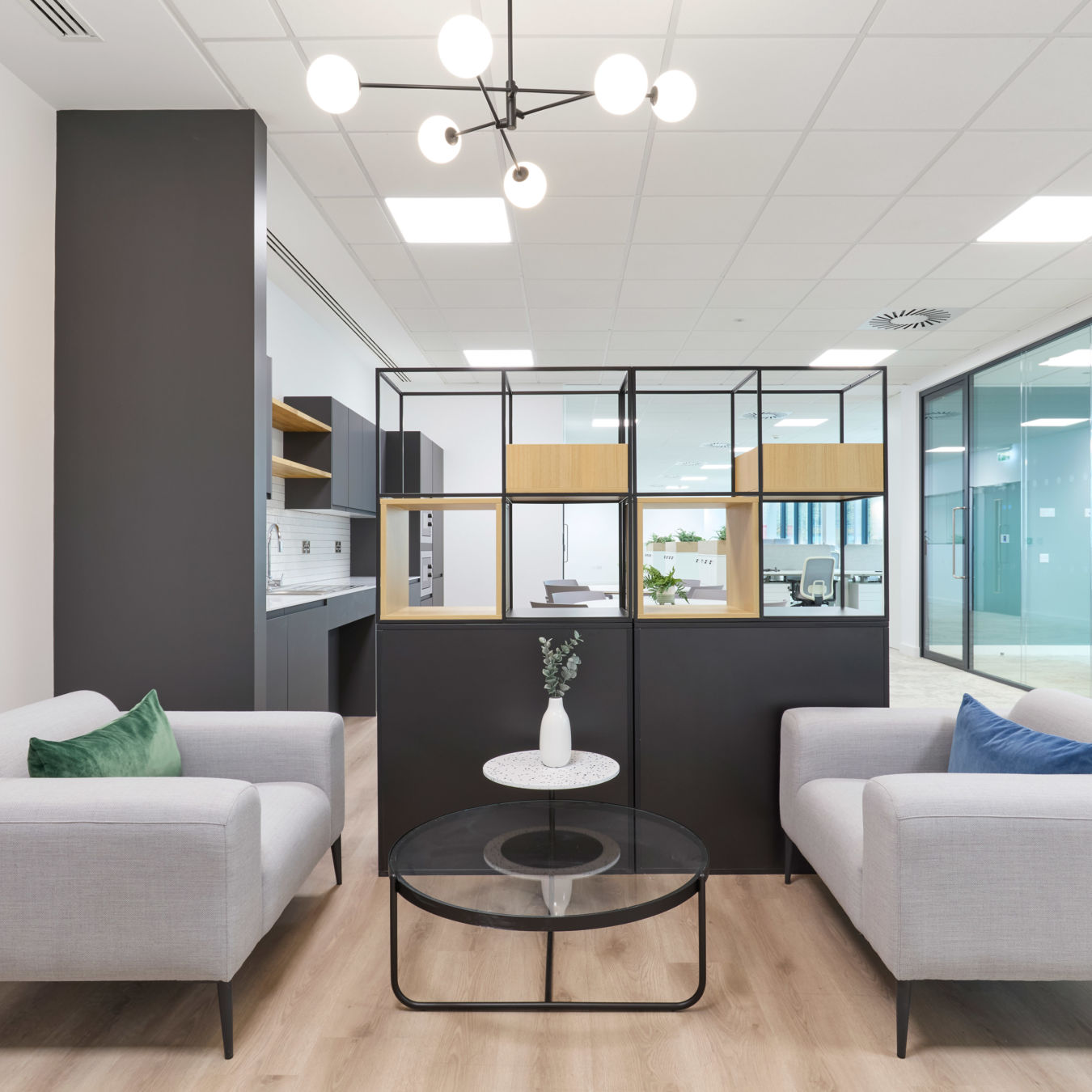 Modern office lounge with two gray sofas facing each other, accented by green and blue pillows. A black grid partition separates the space.