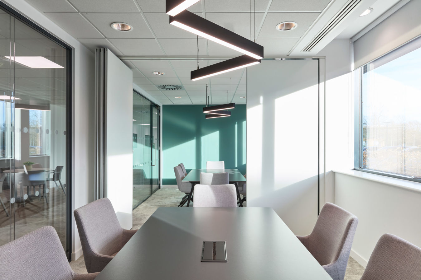 Ceiling lights illuminate a modern conference room with a long table, gray chairs, and a teal accent wall. Glass doors and sunlight create an airy, contemporary atmosphere.