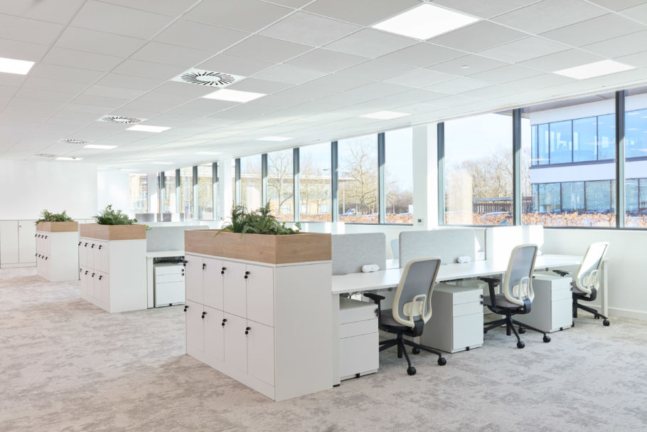 Modern open-plan office space with rows of white desks and swivel chairs. Large windows allow natural light. Planters add greenery, creating a calm atmosphere