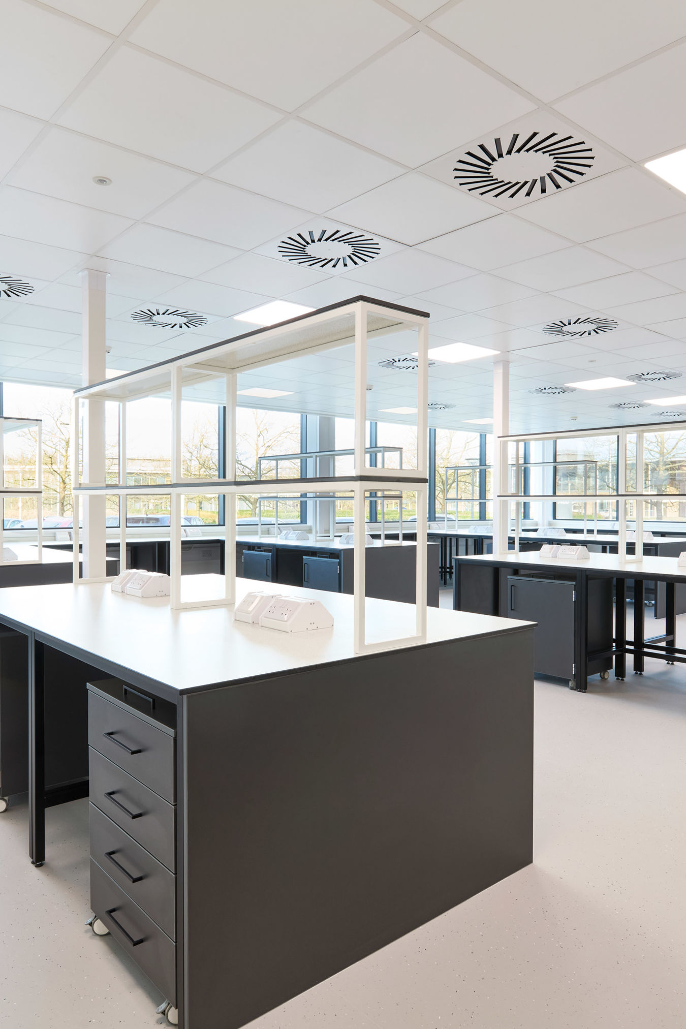 Modern laboratory with black workbenches, white countertops, overhead shelving, and large windows allowing in abundant natural light.