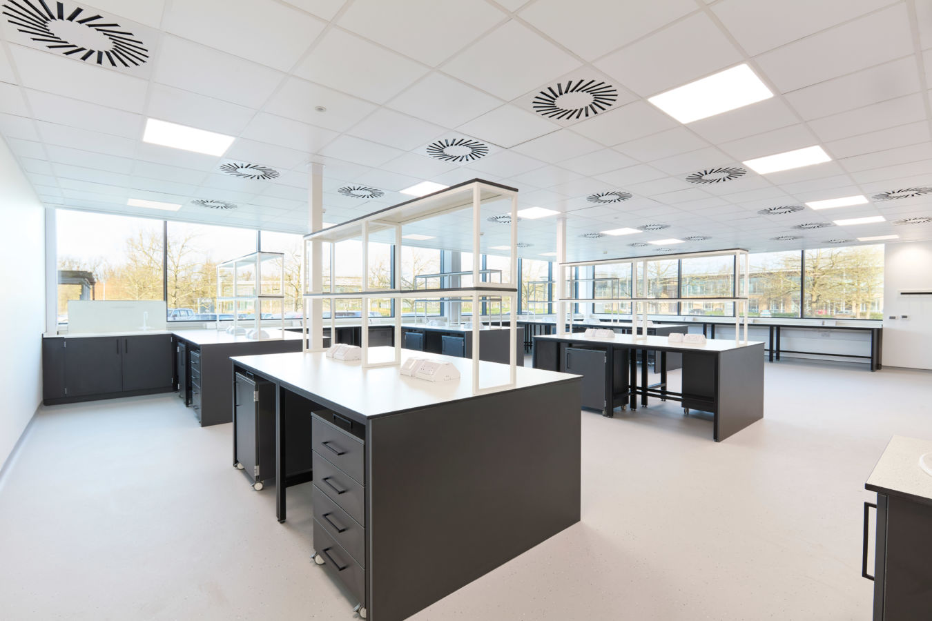 Modern laboratory with black workbenches, white countertops, overhead shelving, and large windows allowing in abundant natural light.