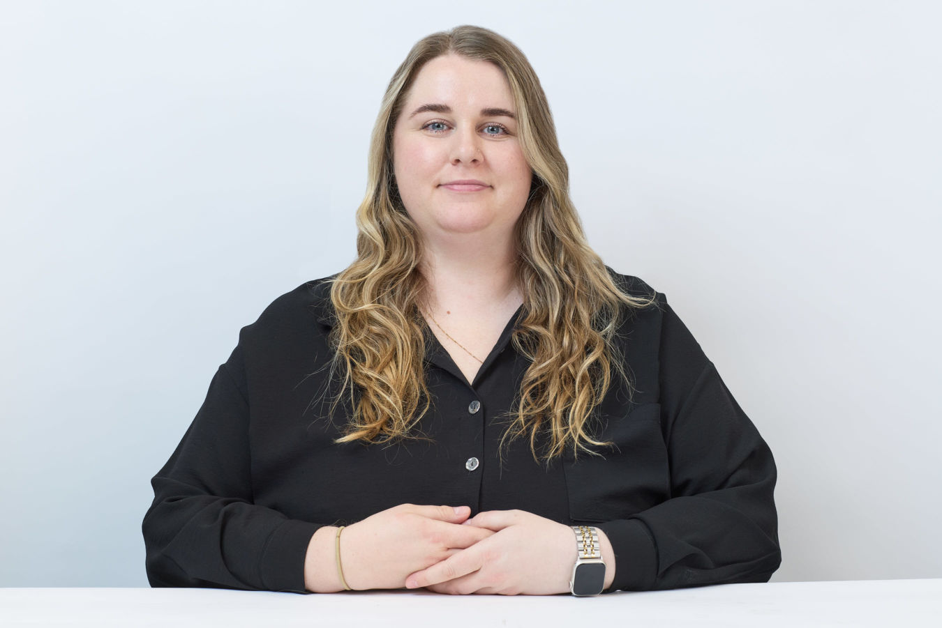A woman with long, wavy hair sits at a white table, wearing a black shirt and smart watch, against a plain light background, hands folded in front of her.