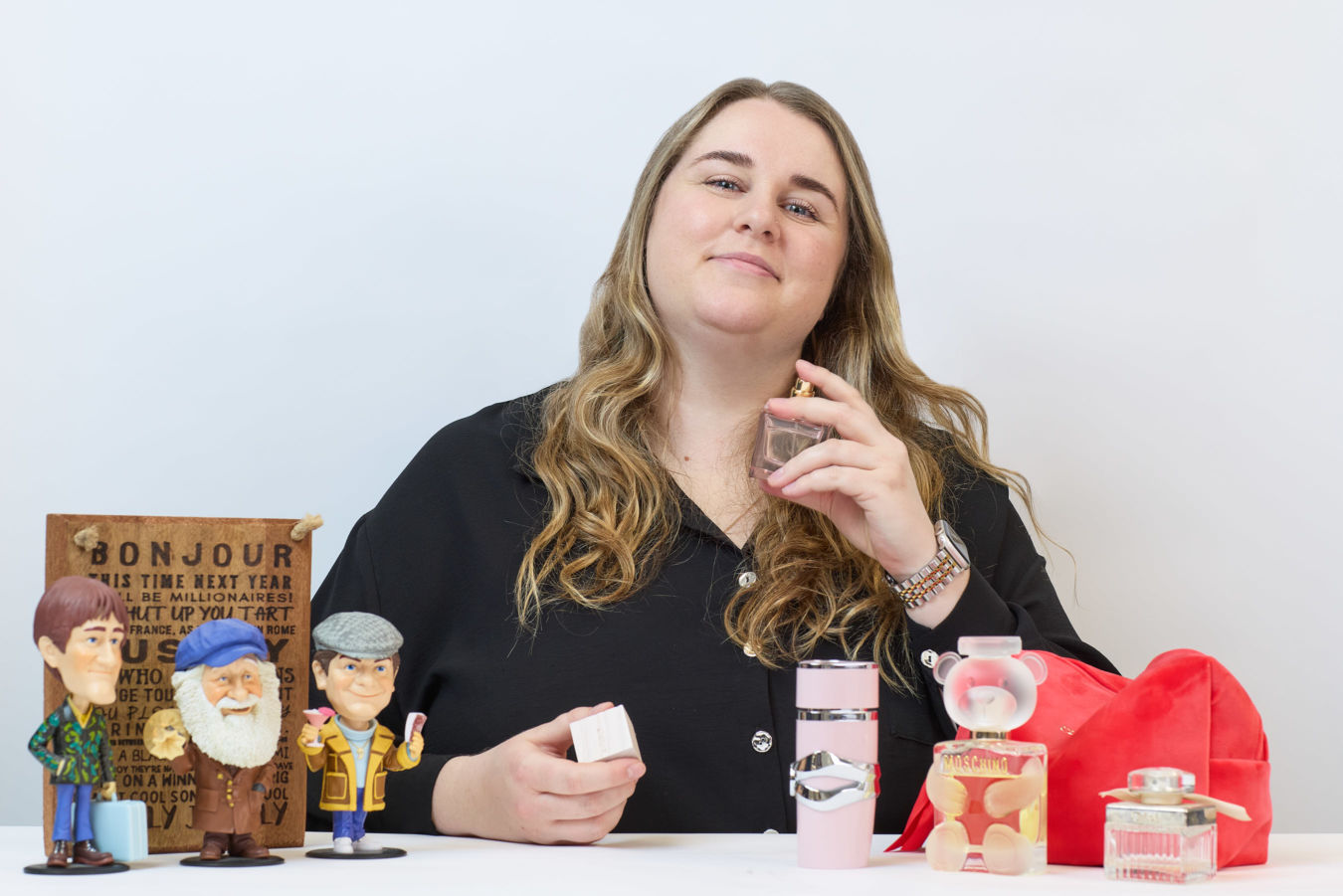 A person holds a perfume bottle near their neck, surrounded by fun figurines and various perfume bottles on a table. A wooden sign in the background reads partially: "BONJOUR THIS TIME NEXT YEAR…"