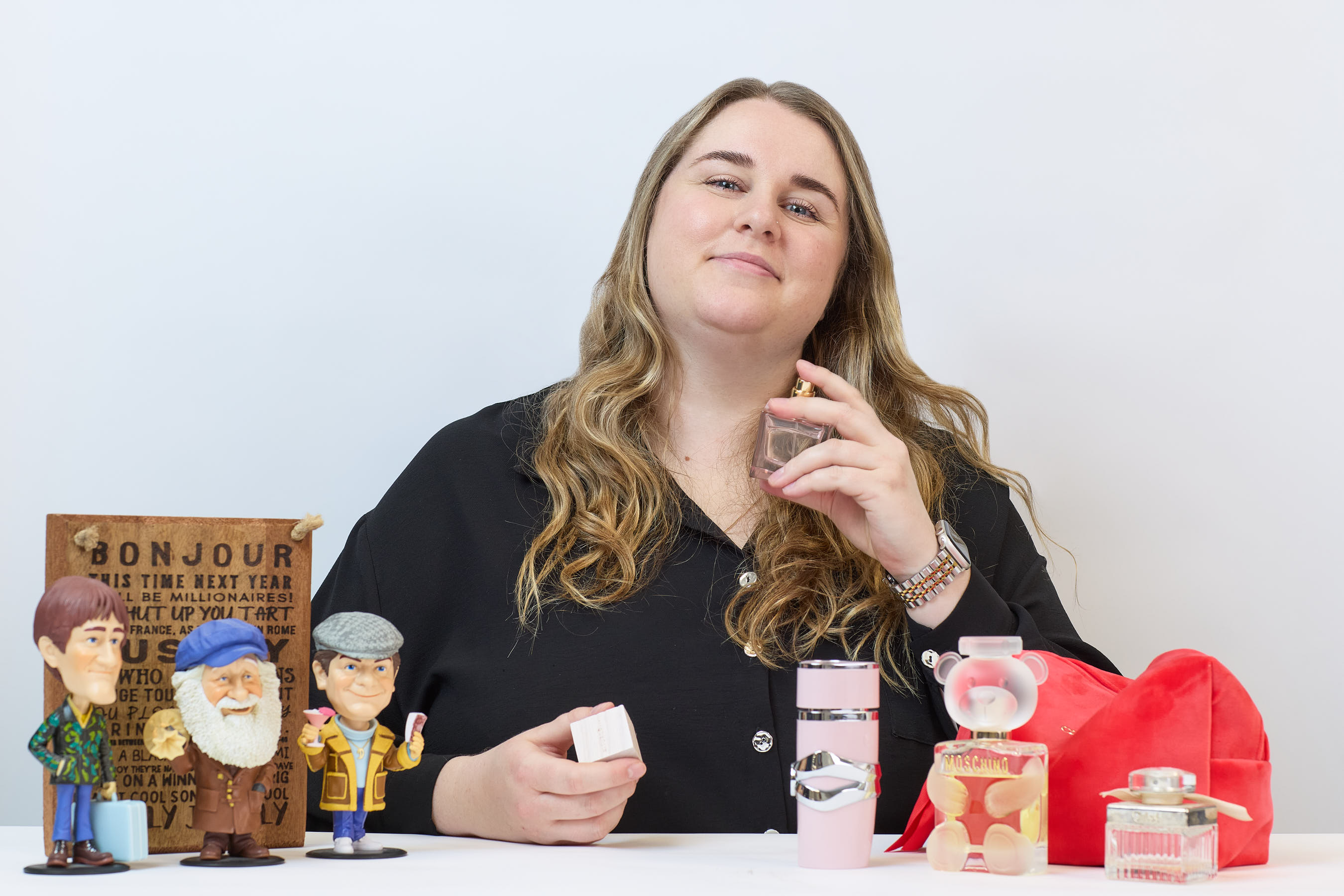 A person holds a perfume bottle near their neck, surrounded by fun figurines and various perfume bottles on a table. A wooden sign in the background reads partially: "BONJOUR THIS TIME NEXT YEAR…"