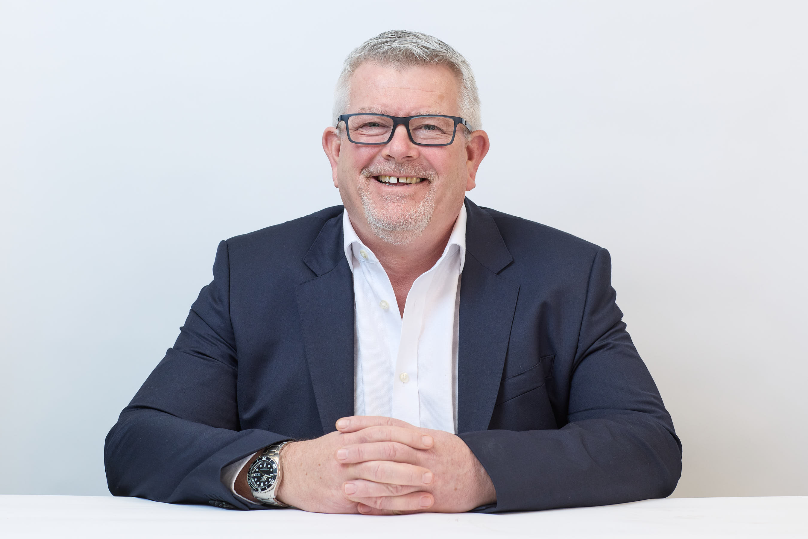 Smiling man in a dark blazer and white shirt, seated with hands clasped on a white table, wearing glasses and a wristwatch, against a plain white background.