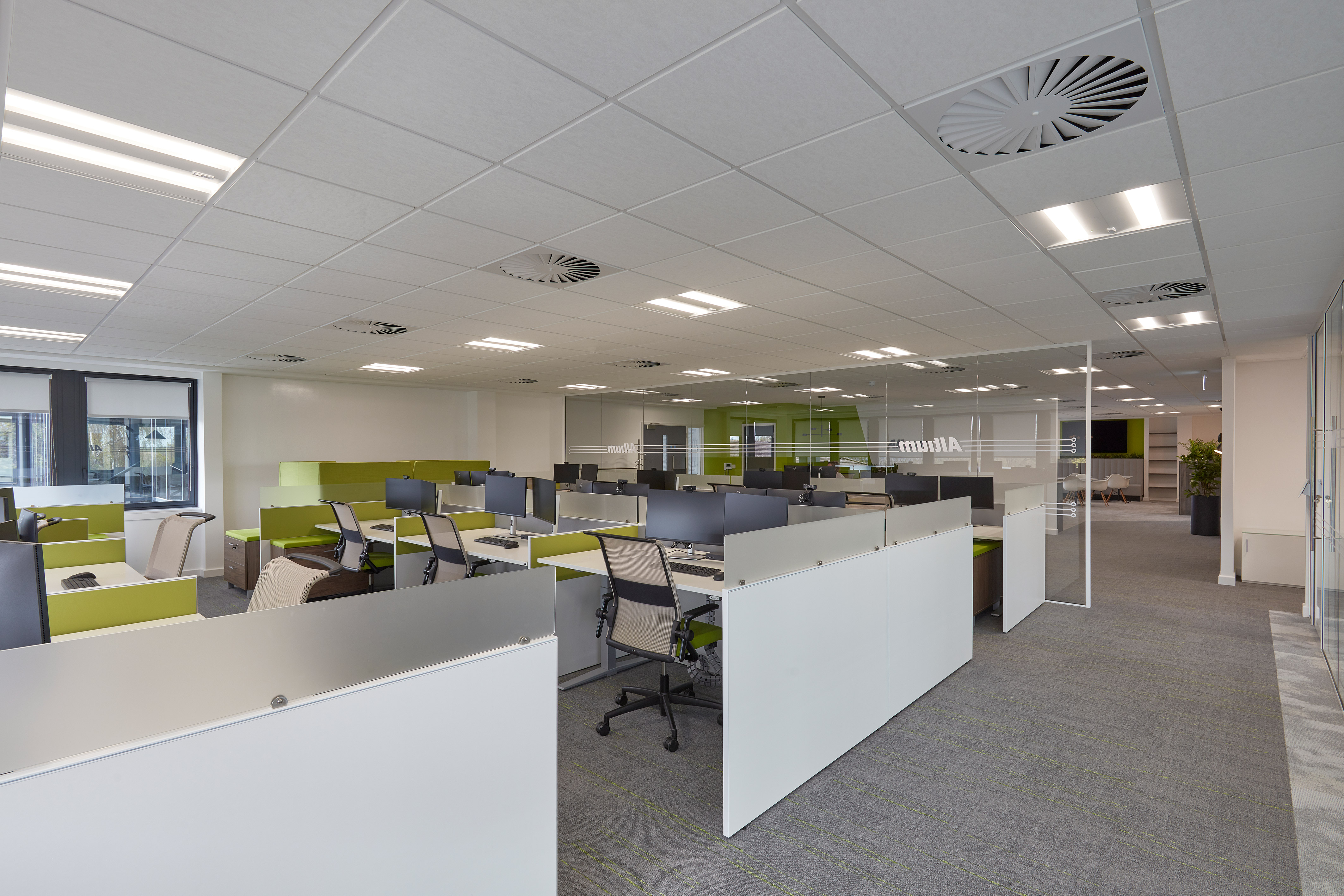 Open-plan office with green and white workstations, ergonomic chairs, and computers, in a spacious area with ceiling lights, glass dividers, and the word "Altium" visible on a partition.