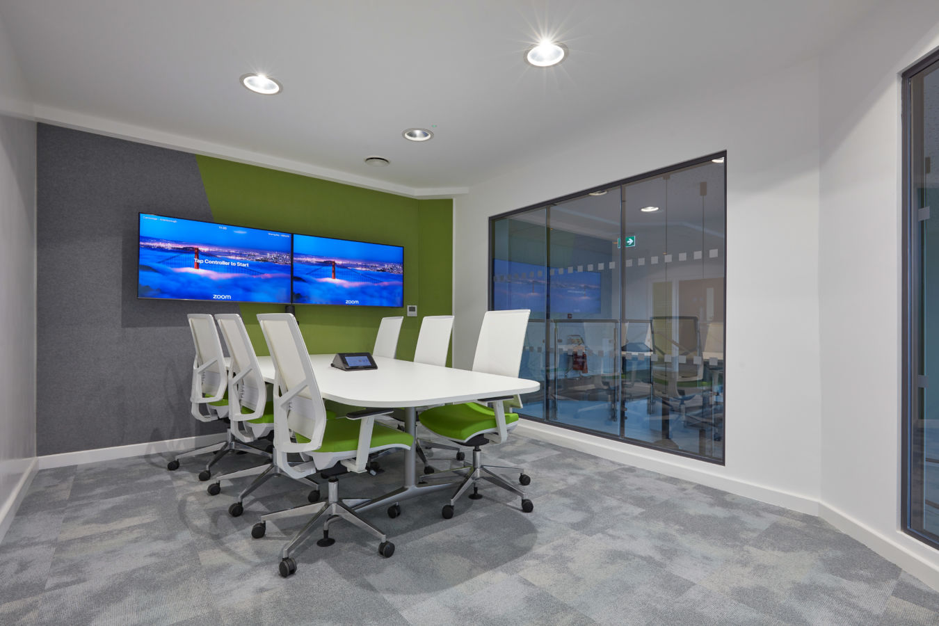 Conference room featuring a white oval table with six white chairs on wheels, facing two wall-mounted screens displaying "Zoom." A large interior glass window separates the space.