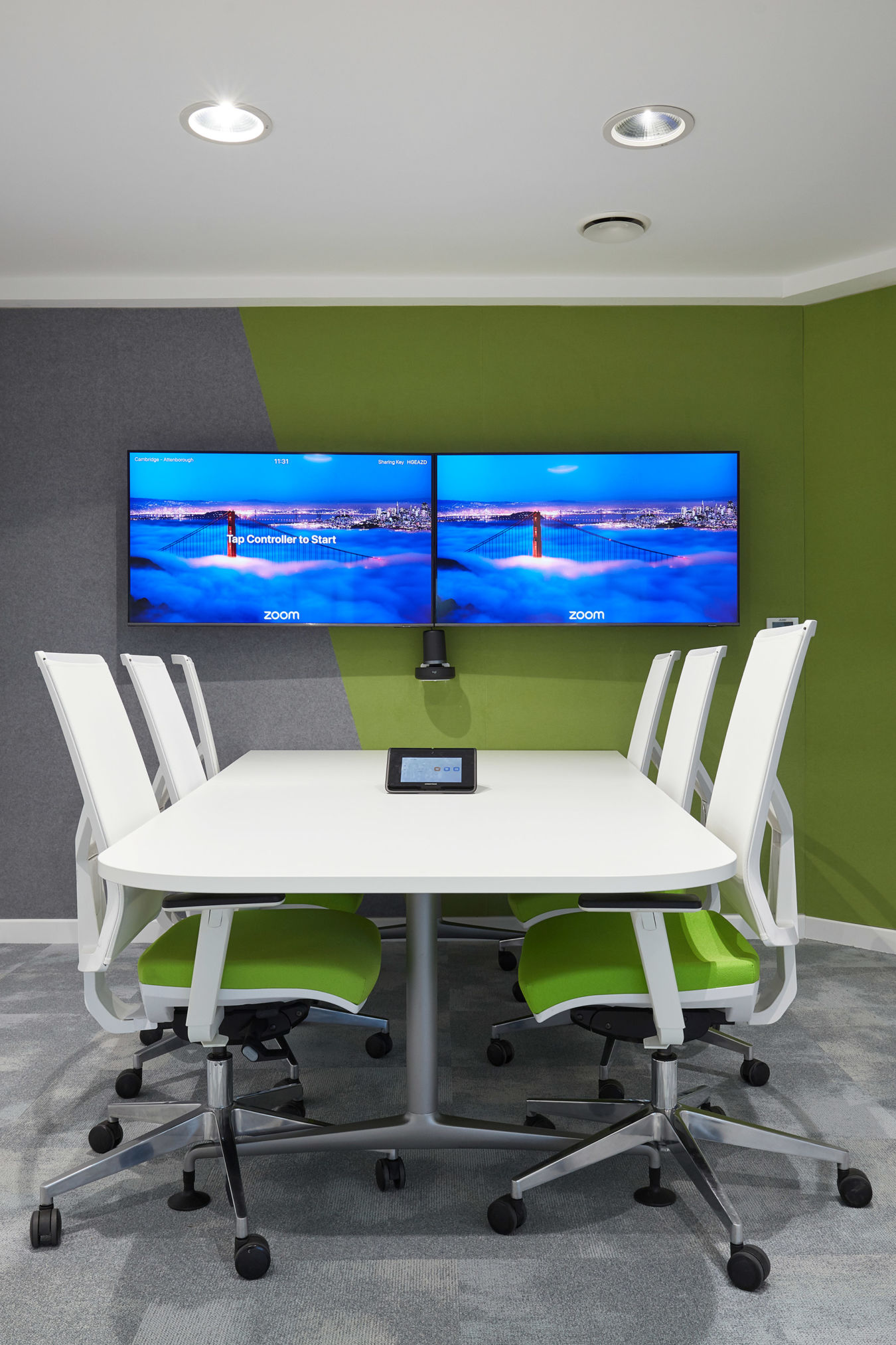 Modern conference room with green accent walls, two large screens, and a white table surrounded by white chairs with green seats. Glass wall on the righ