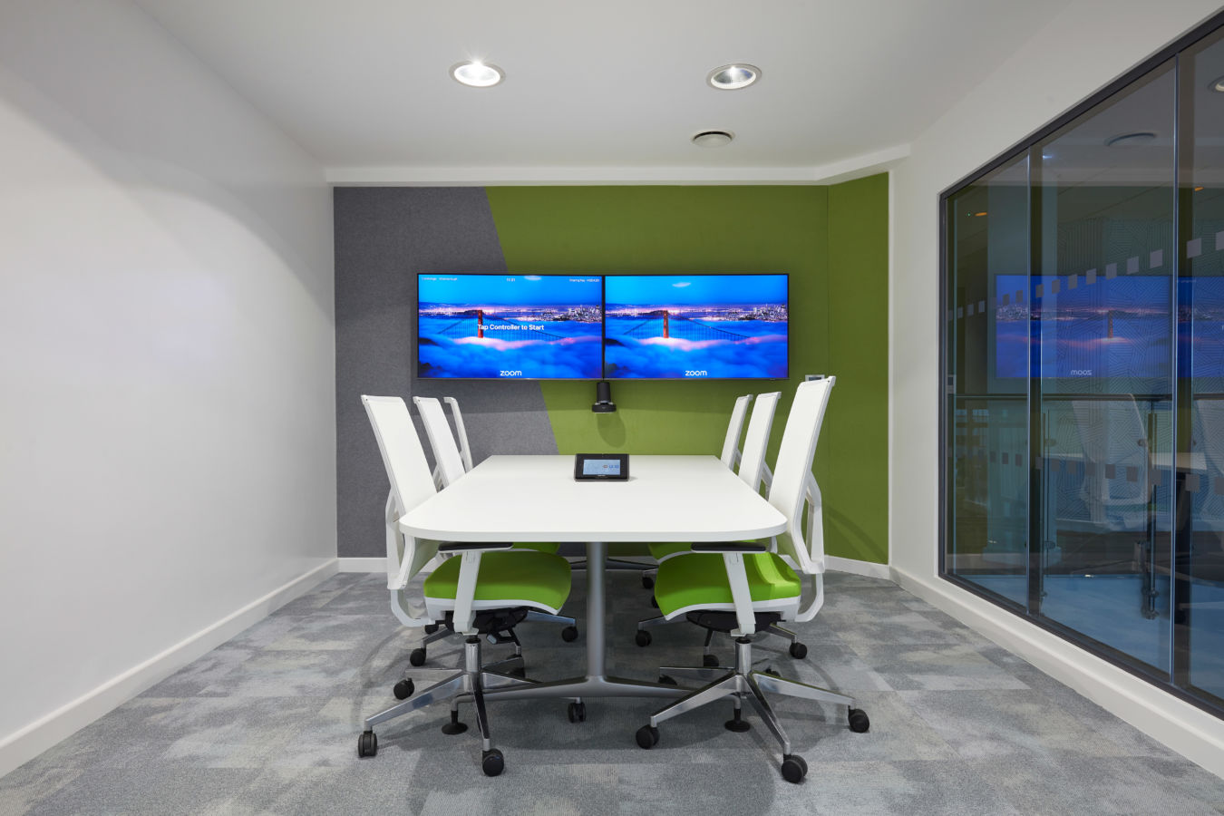 Modern conference room with green accent walls, two large screens, and a white table surrounded by white chairs with green seats. Glass wall on the righ
