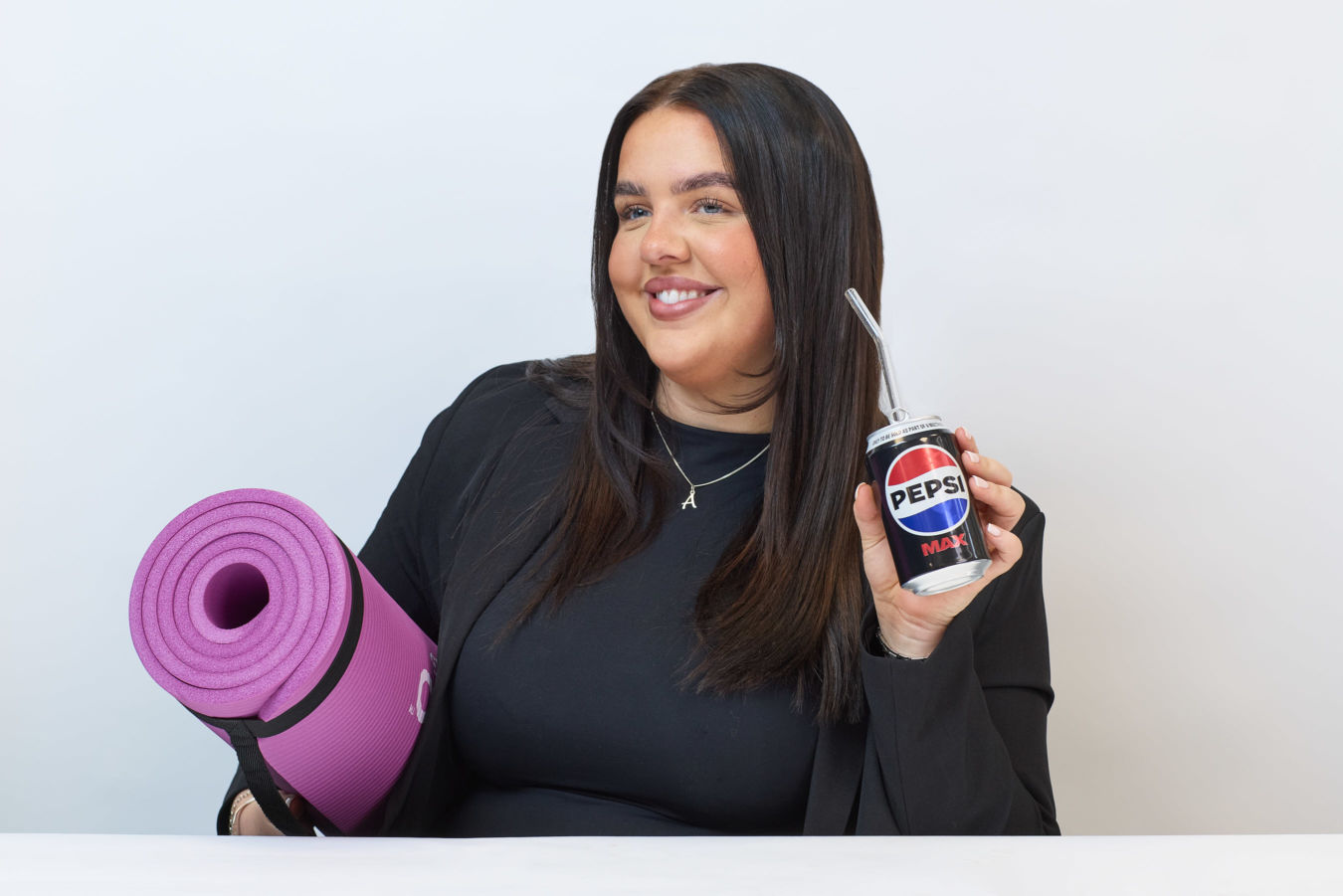 A woman holds a rolled purple yoga mat and a Pepsi Max can with a straw, smiling against a plain white background.