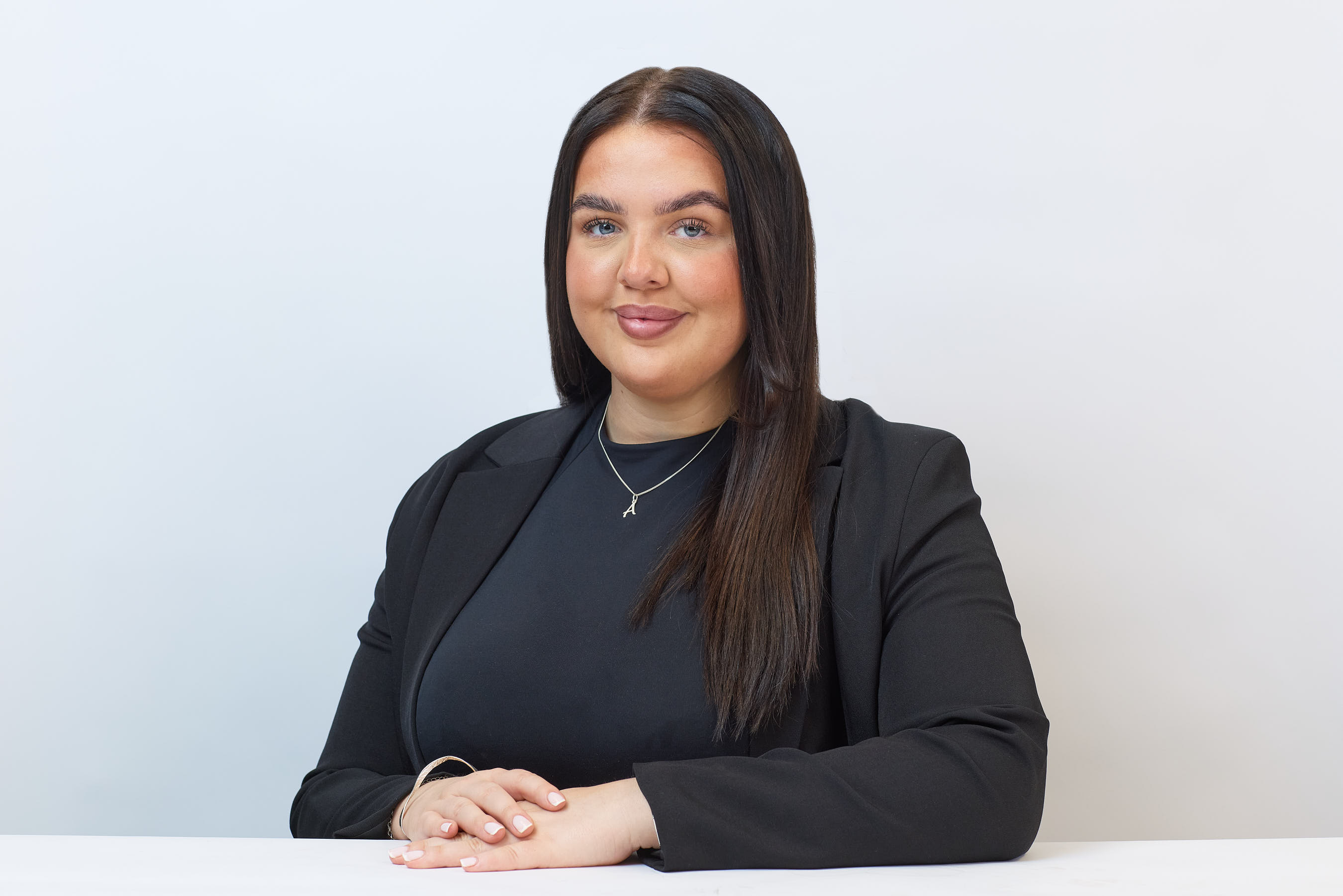 A person with long dark hair, wearing a black blazer and necklace, sits with hands folded, smiling against a white background.