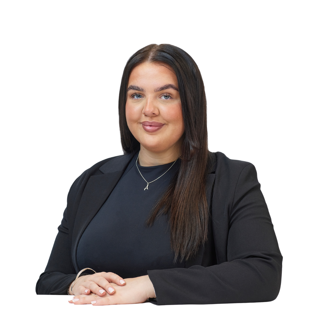 A woman with long brown hair and a black blazer sits at a table, hands folded, against a plain white background. She wears a necklace with the letter "A".