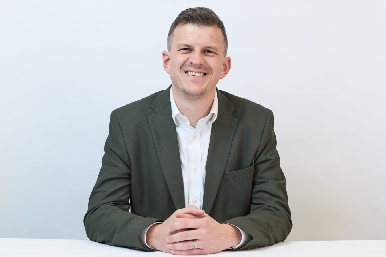 A person in a green suit and white shirt smiles while sitting at a white table against a plain white background.