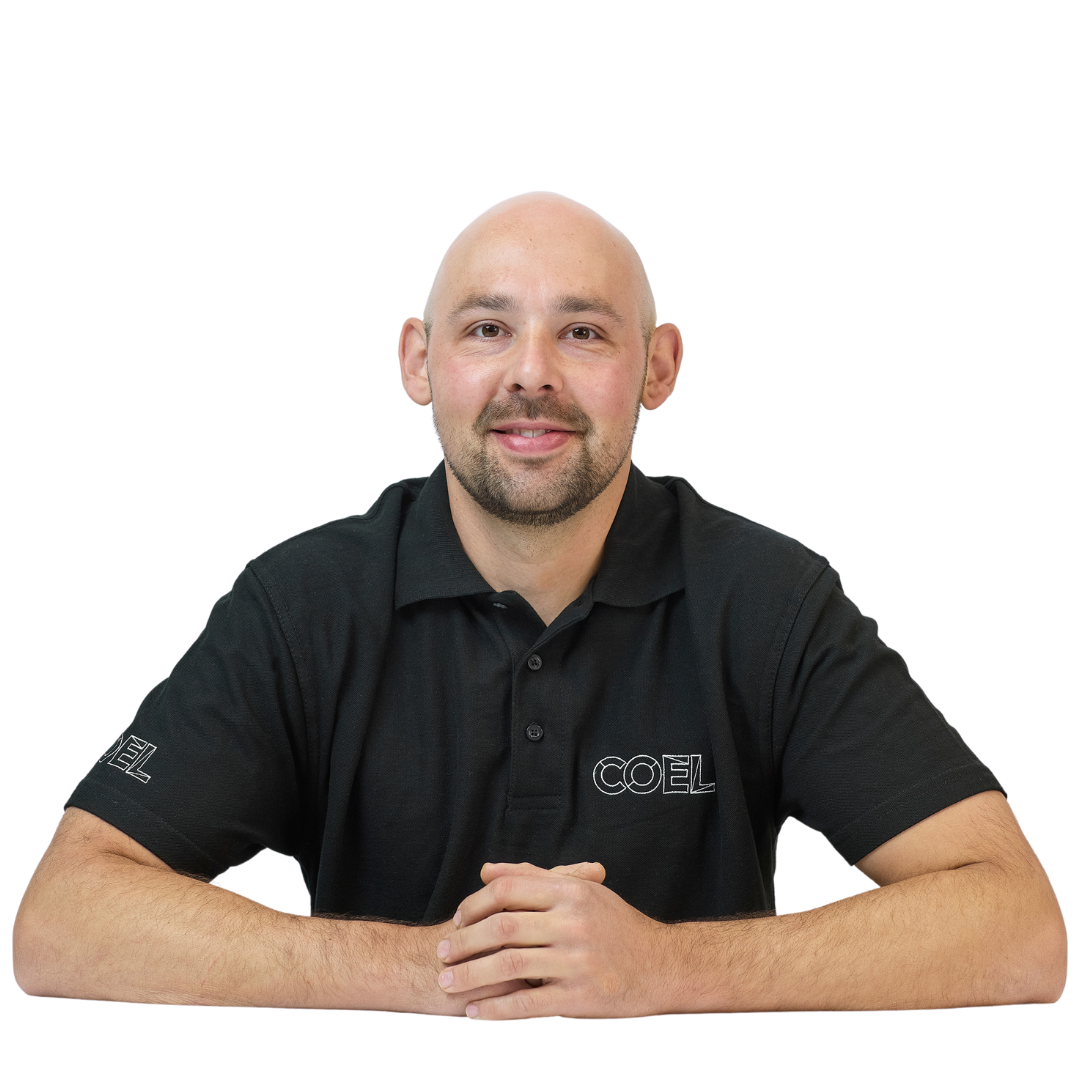A person sits with hands clasped on a table, wearing a black polo shirt with "COEL" on the chest and sleeve, against a plain white background.