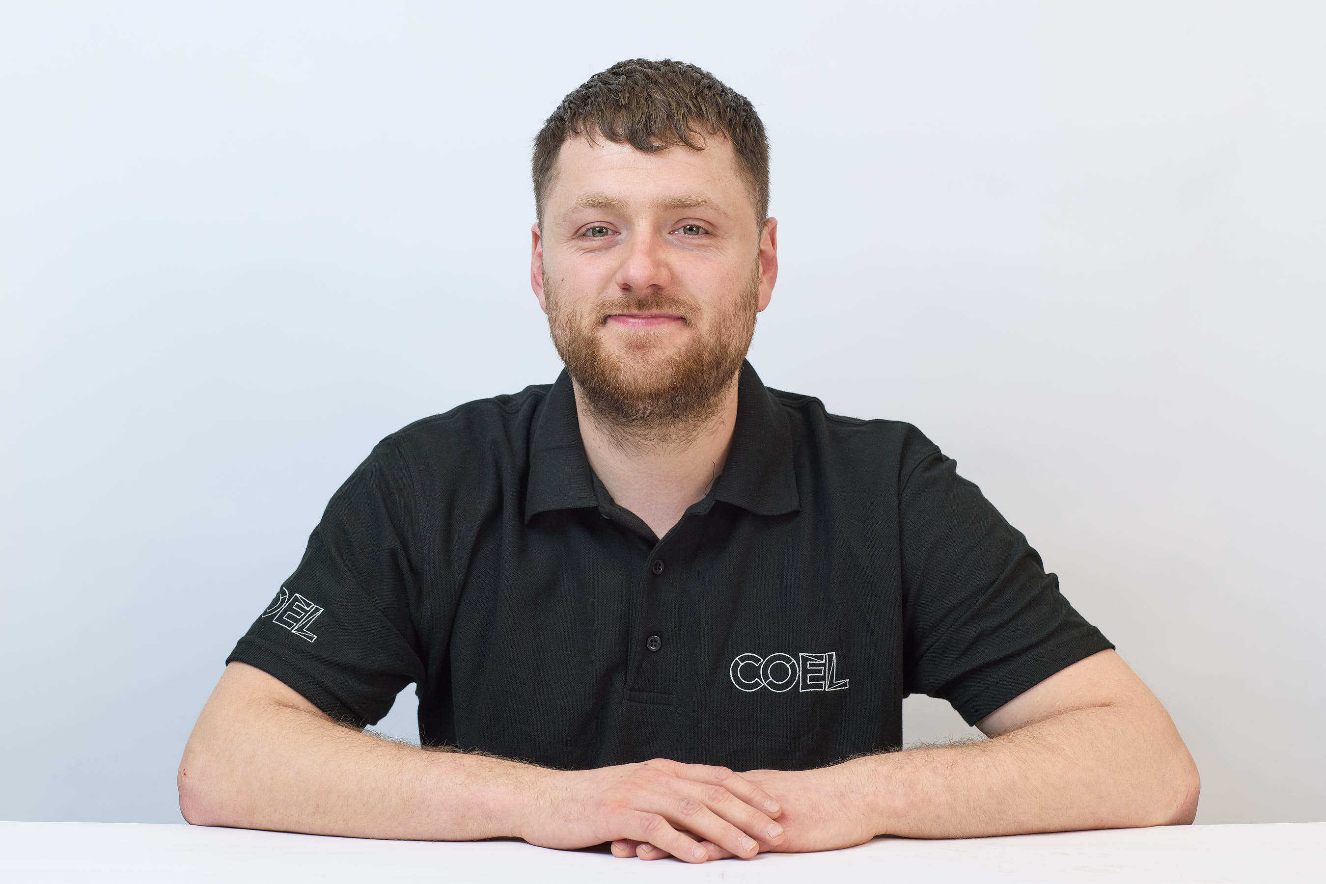 A person wearing a black polo shirt with "COEL" text, seated at a white table, against a plain white background.