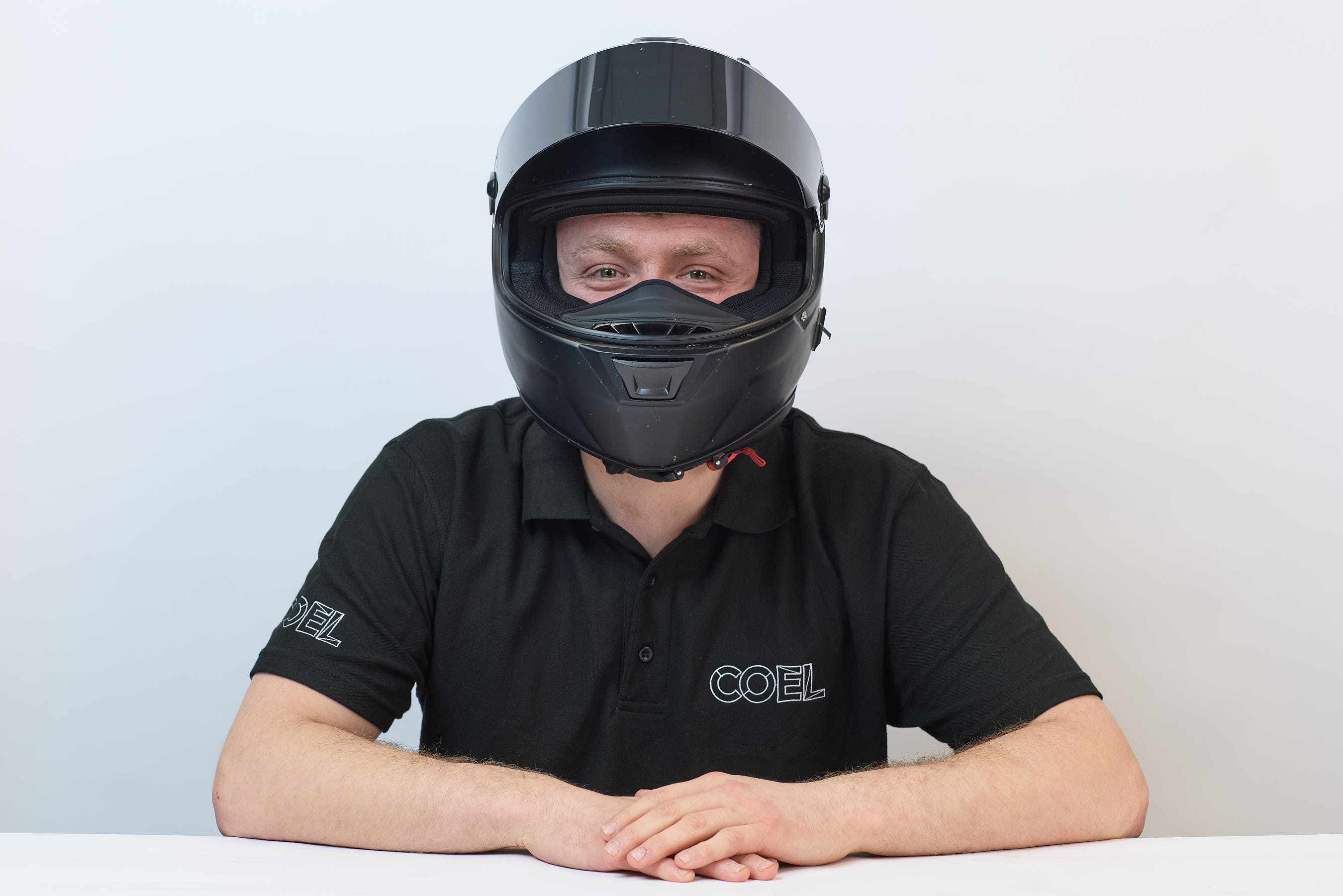A person wearing a black motorcycle helmet and a black shirt with "COEL" embroidered on it sits against a plain white background.