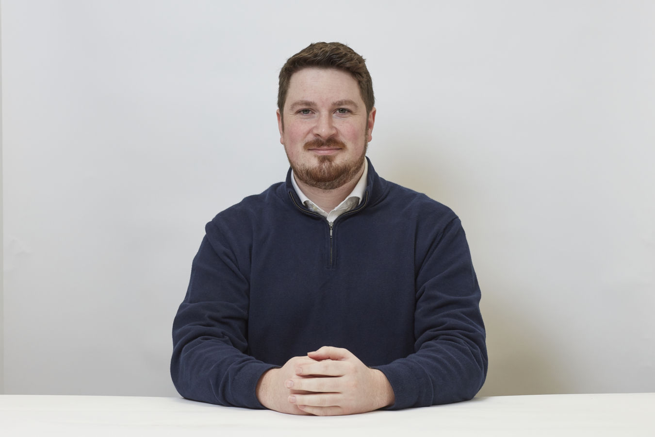 A man with short hair and a beard sits at a table, wearing a navy sweater, hands clasped, against a plain white background.