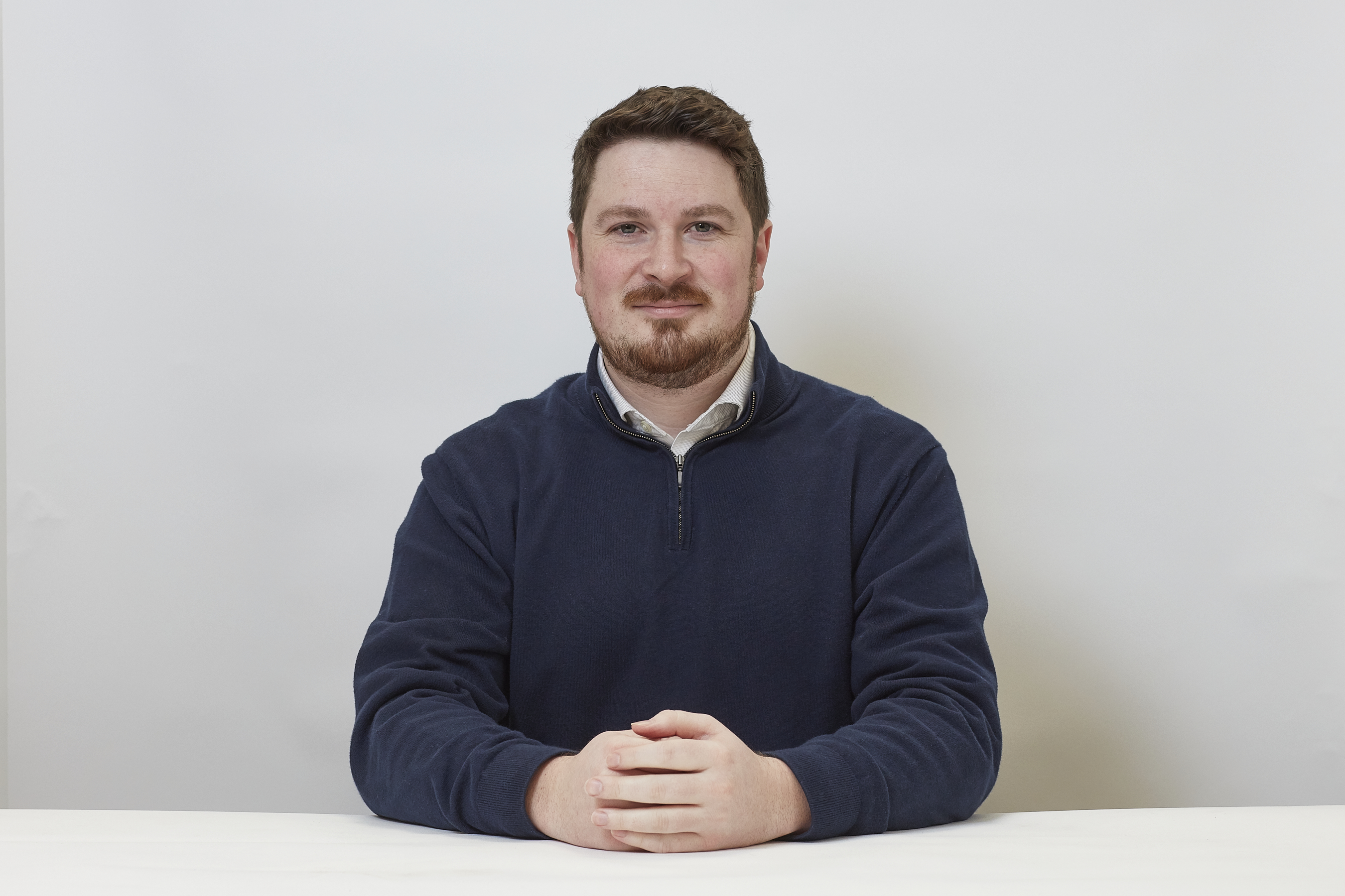 A man with short hair and a beard sits at a table, wearing a navy sweater, hands clasped, against a plain white background.