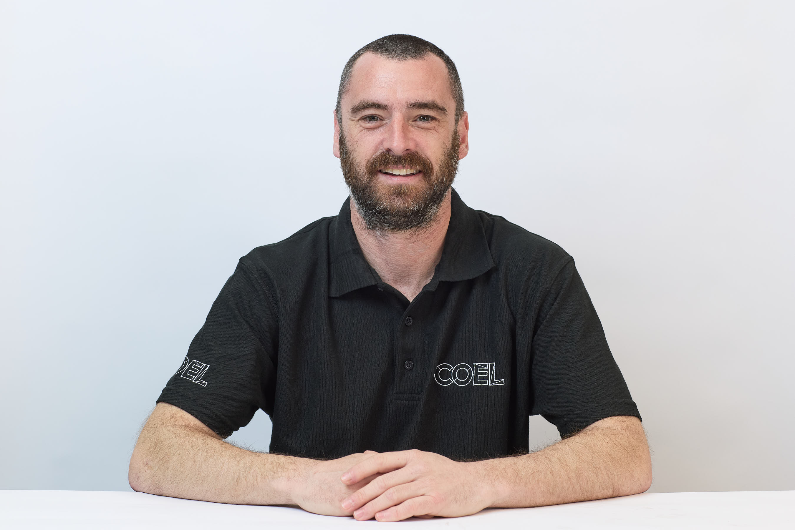 A man with a beard smiles, wearing a black polo shirt with "COEL" printed on it, sitting against a plain white background.