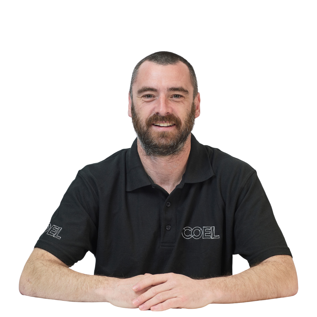 Man with a beard smiles while sitting at a table, wearing a black polo shirt with "COEL" text on it, against a plain white background.