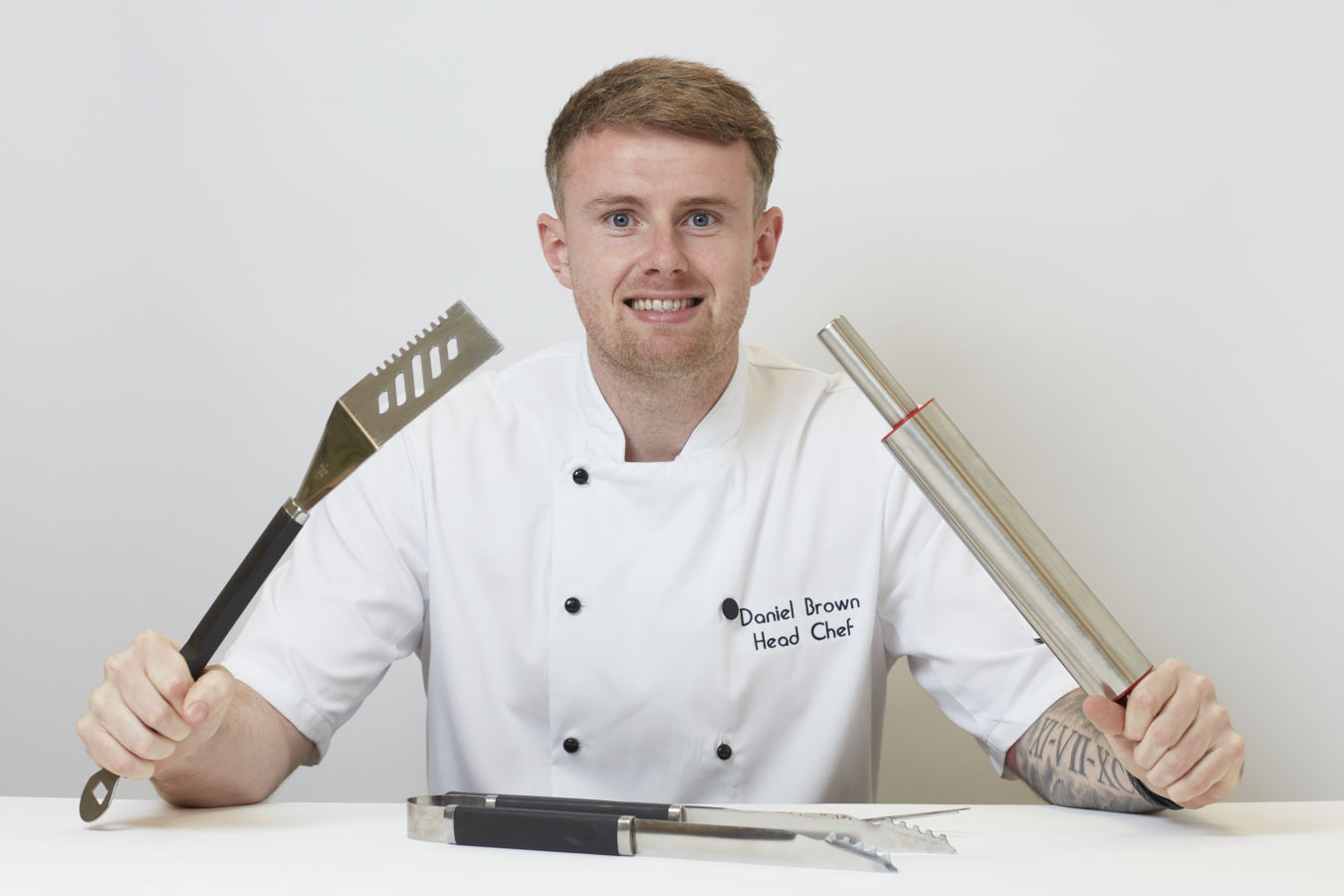 Chef holding spatula and rolling pin, smiling at camera in a white uniform with "Daniel Brown Head Chef" text, sitting at a table with tongs on a plain background.