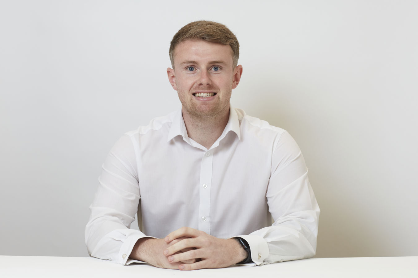 A man in a white shirt sits with hands folded on a table, smiling against a plain white background.