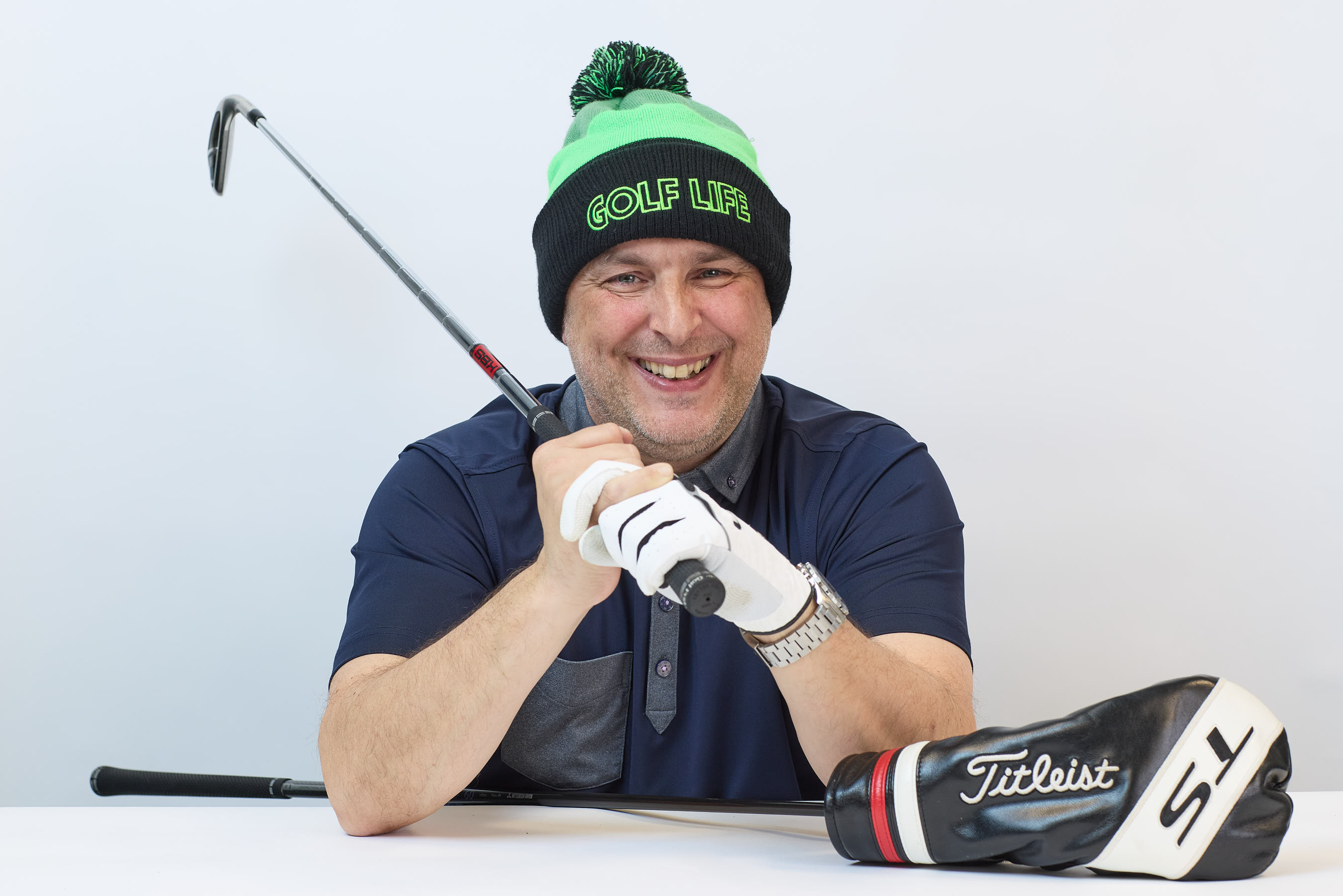 Smiling man wearing a "GOLF LIFE" beanie holds a golf club across his shoulder, with a Titleist driver headcover on a table in a bright setting.