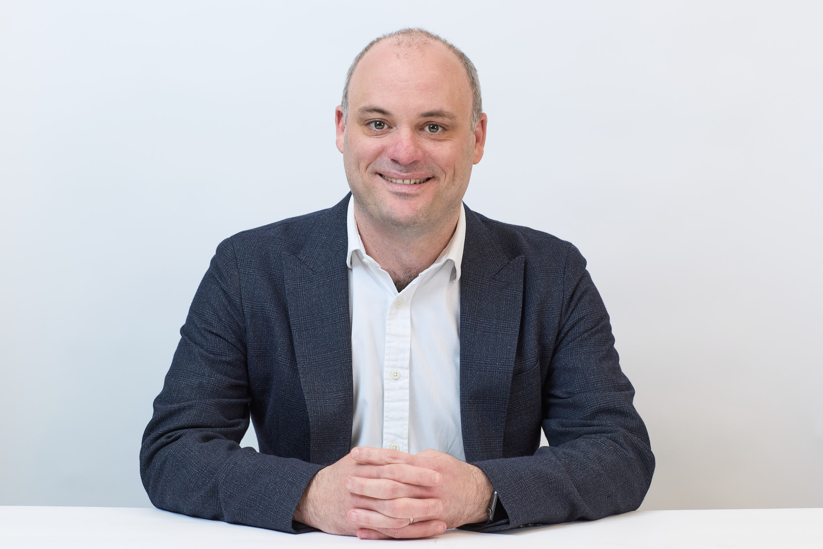 Smiling person sits at a white table with hands clasped, wearing a dark blazer and white shirt, against a plain light background.