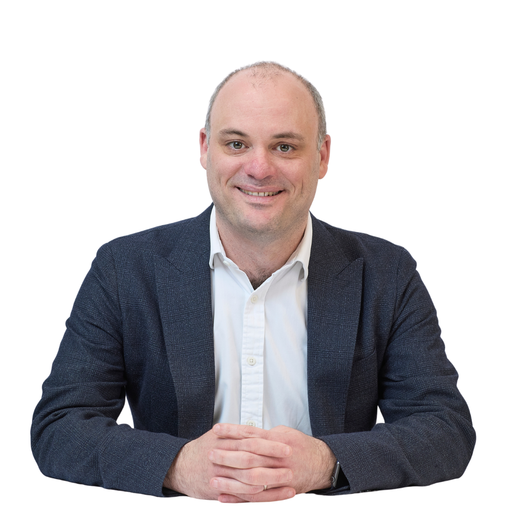 A person in a dark blazer and white shirt sits at a table, hands clasped, smiling. The background is plain white.
