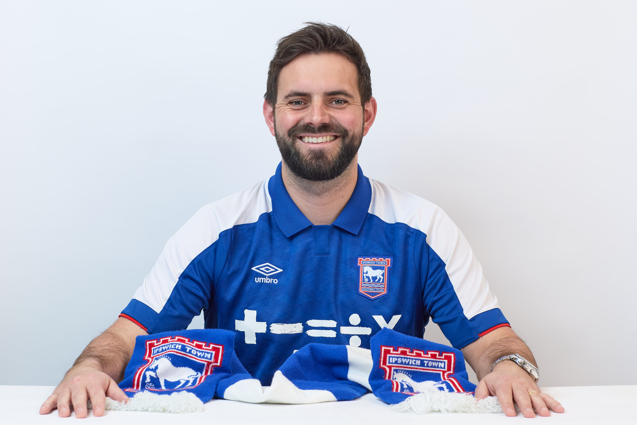 A man wearing a blue Ipswich Town FC shirt smiles, sitting behind a matching scarf with the text "IPSWICH TOWN" on a white background.