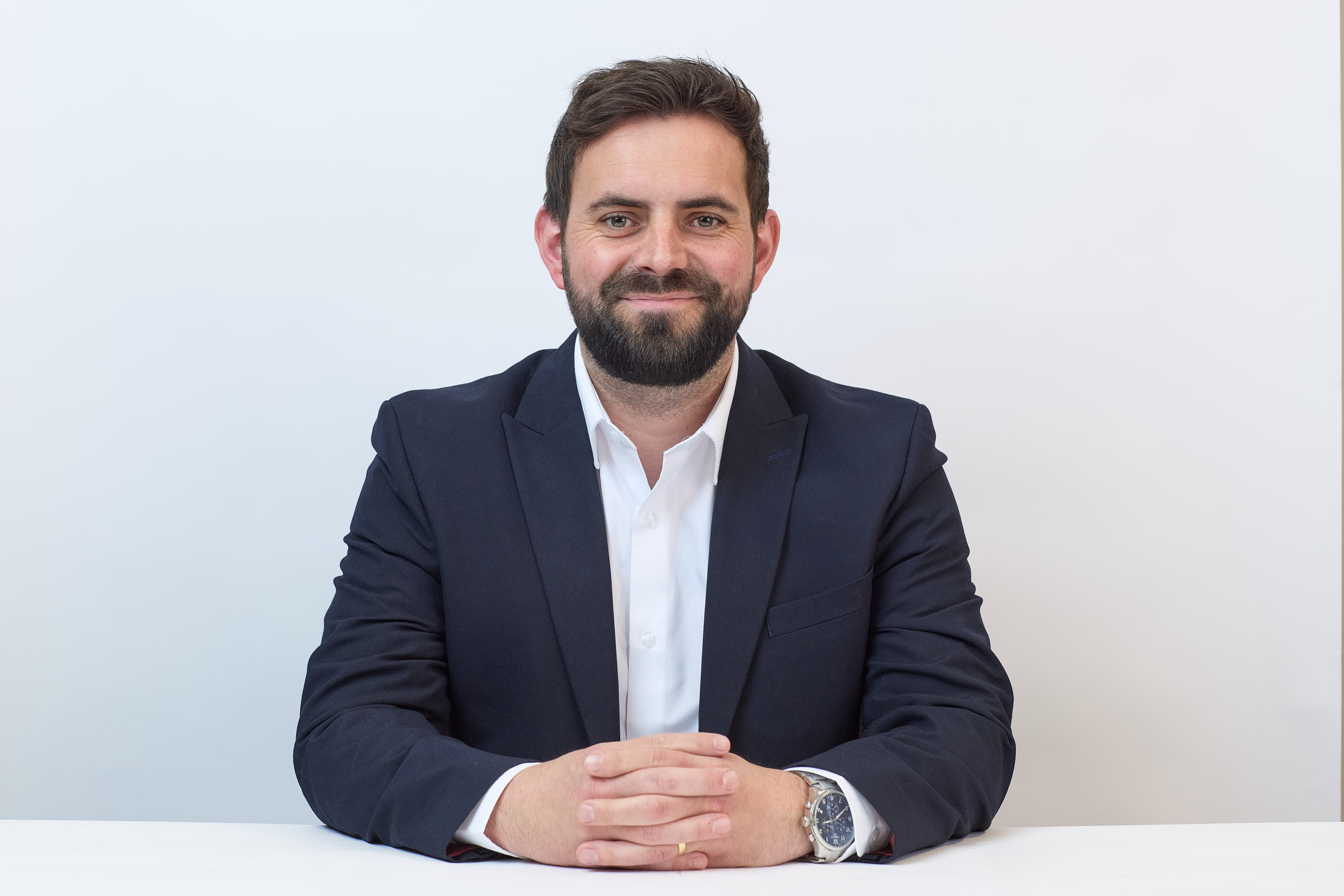 A bearded man in a navy suit sits with folded hands, smiling confidently against a white background.