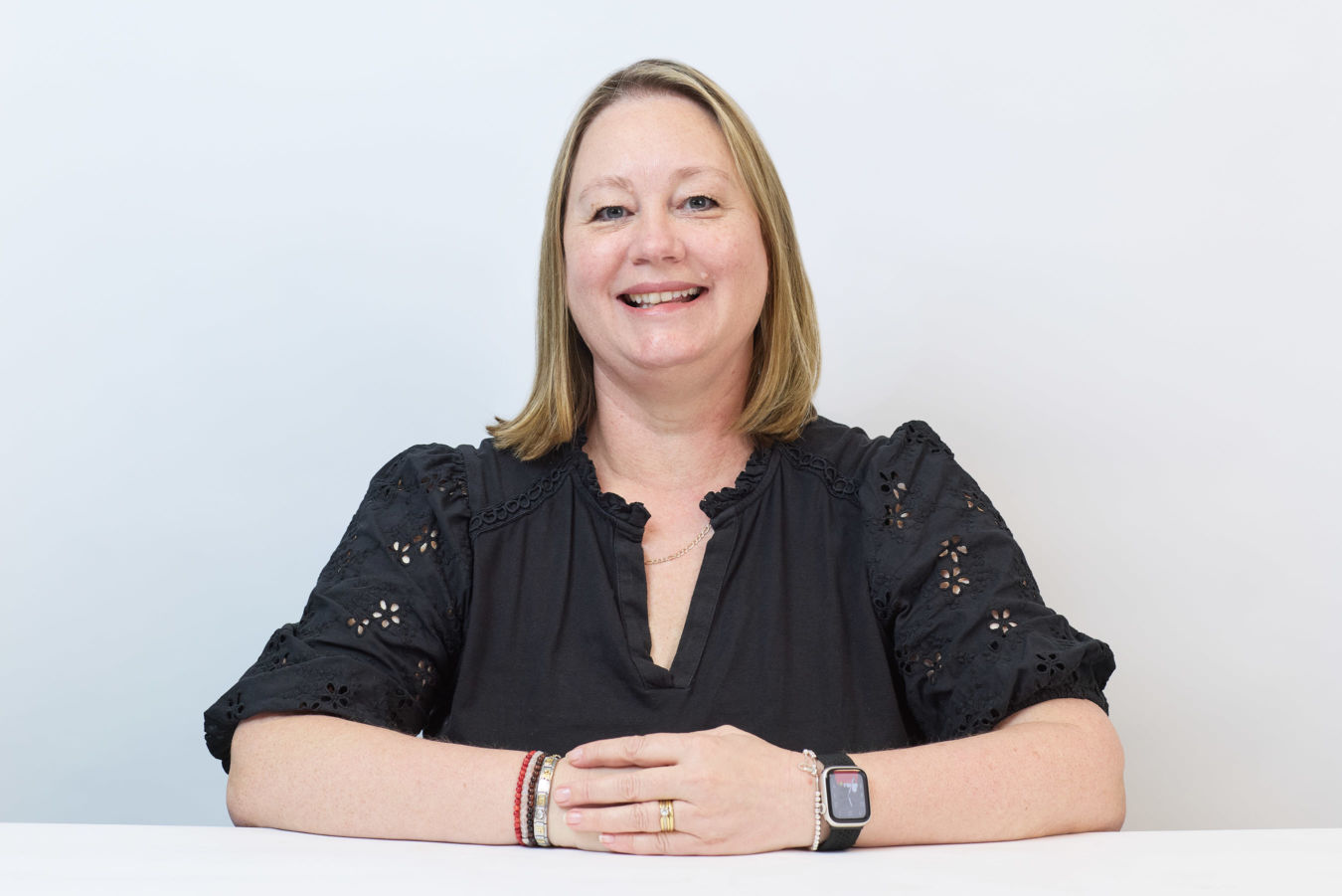 A woman with shoulder-length blonde hair is smiling, resting her hands on a table, wearing a black blouse and a smartwatch, against a plain white background.