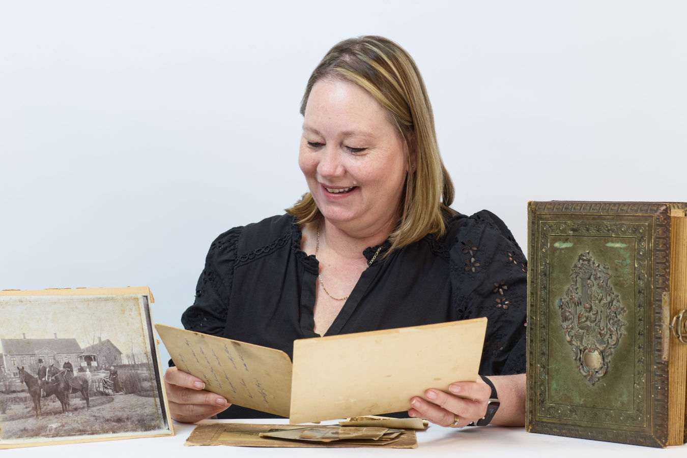 A woman, smiling and seated, examines a vintage document in a bright setting. Around her are an antique photo of people with horses and an ornate old book.