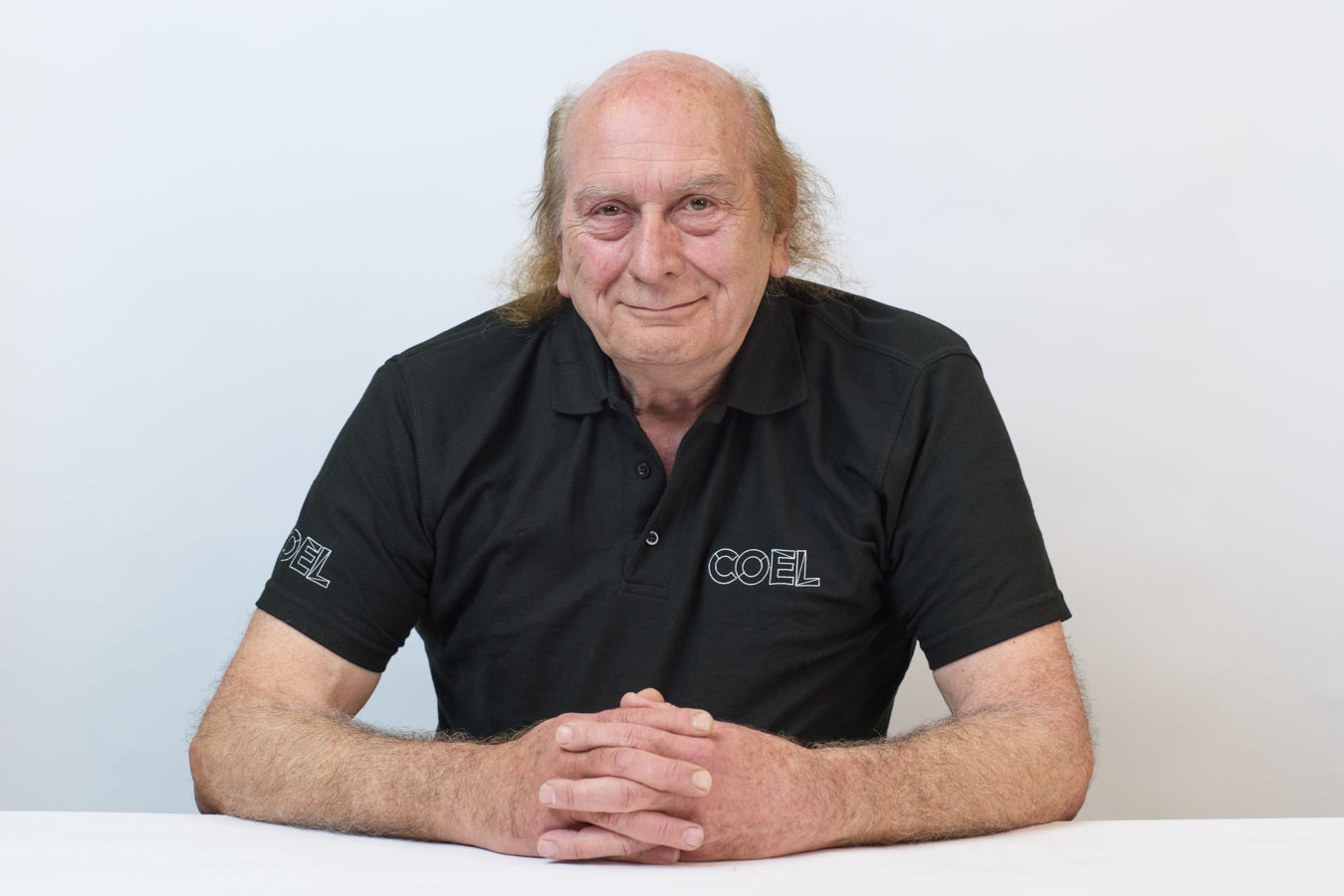 An older man sits at a white table, resting his hands together, wearing a black polo shirt with "COEL" embroidered on it, against a plain white background.