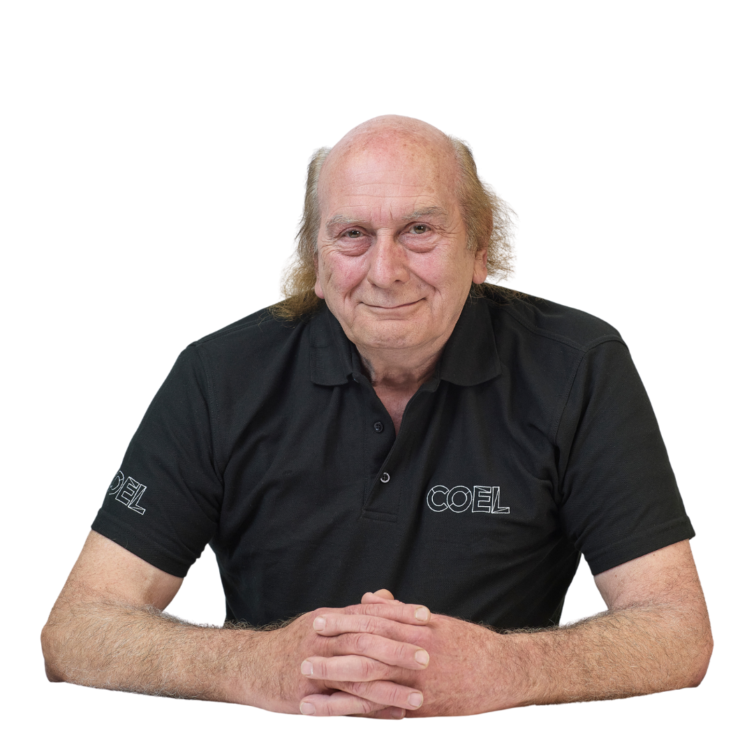 Man wearing a black polo shirt with "COEL" text, sits with hands clasped on a table, smiling against a white background.