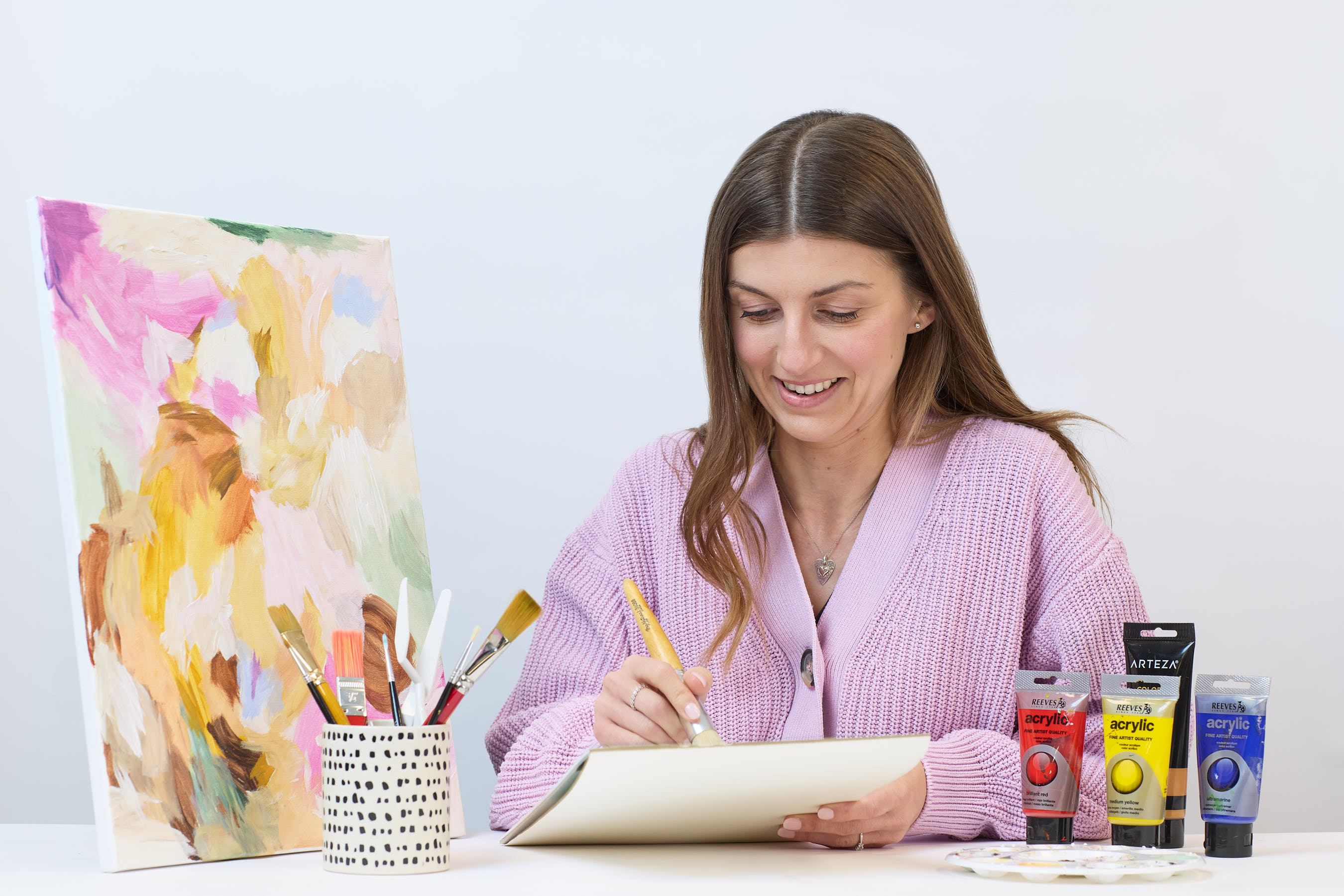 A woman in a pink sweater draws on paper at a table with acrylic paints, brushes in a holder, and an abstract painting.