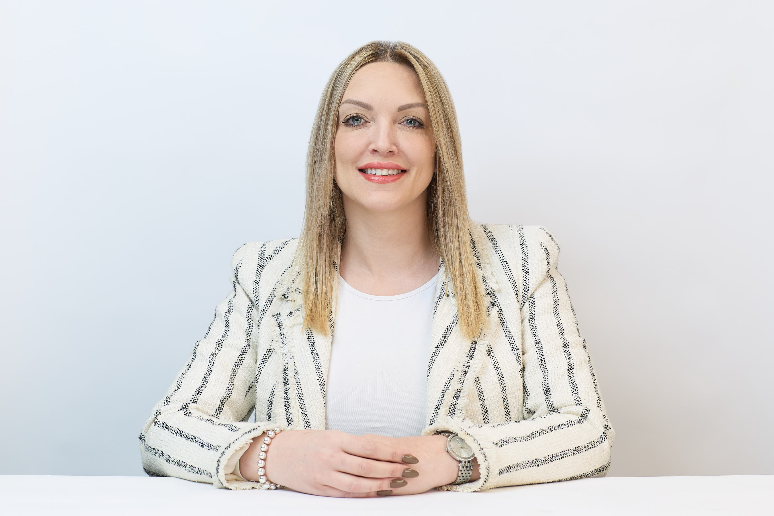 A woman with long blonde hair sits smiling, hands resting on a table. She wears a white striped blazer and a wristwatch, set against a plain white background.