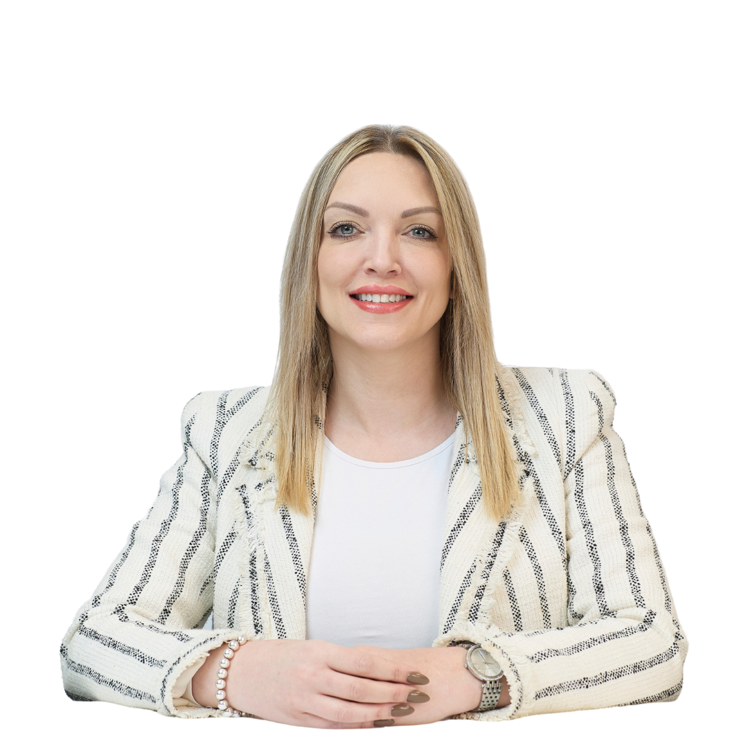 A woman with straight blond hair smiles while seated at a white table, wearing a striped blazer and accessories, against a plain white background.