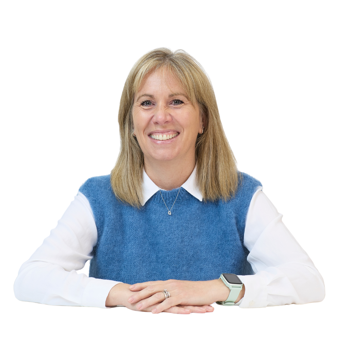A woman wearing a blue sweater vest over a white shirt, smiling with hands clasped on a table, against a plain background. She wears a smartwatch and a small pendant necklace.