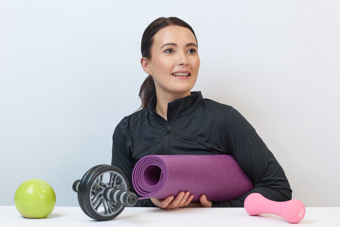 A woman holds a purple yoga mat, smiling, seated at a table with a green ball, ab roller, and pink dumbbell on it, set against a plain background.