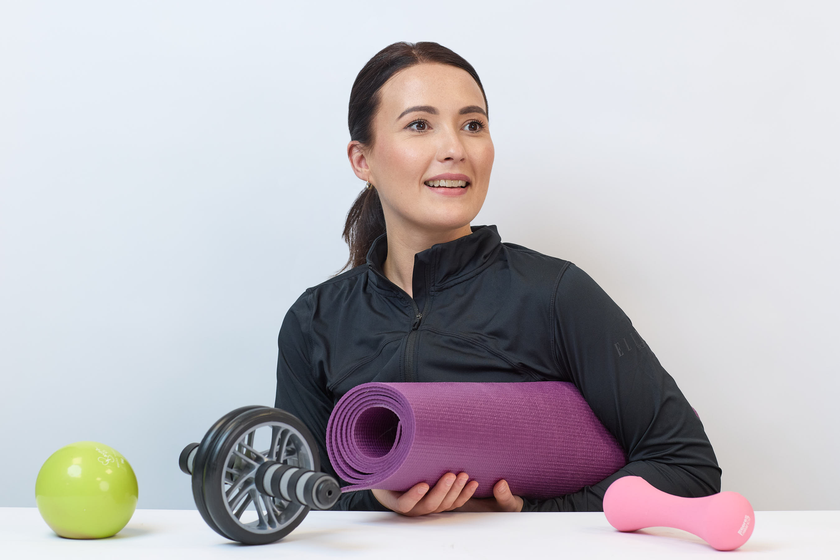 A woman holds a purple yoga mat, smiling, seated at a table with a green ball, ab roller, and pink dumbbell on it, set against a plain background.