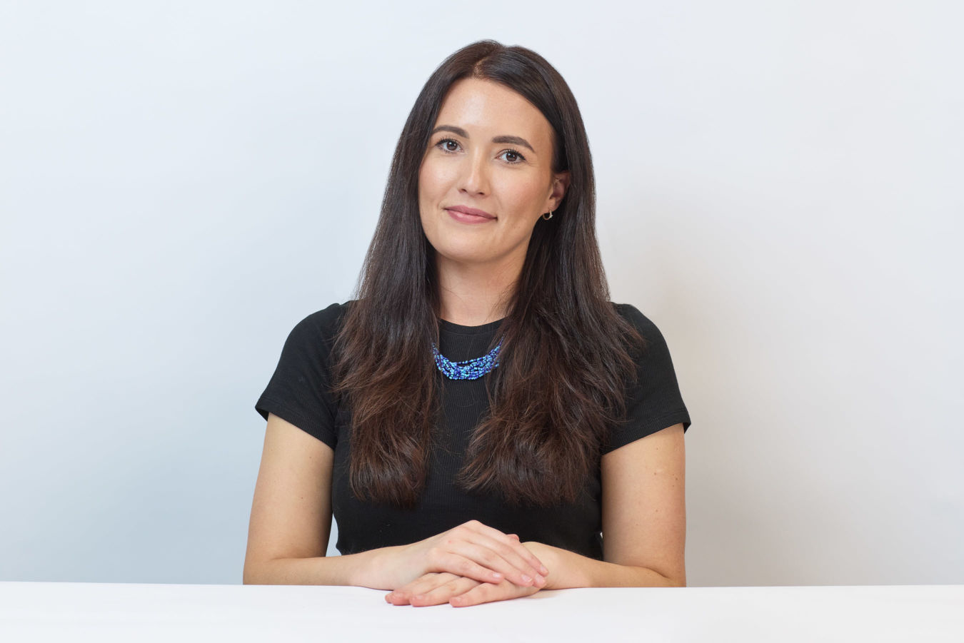 A woman with long dark hair sits at a white table, wearing a black shirt and blue necklace, against a plain white background.
