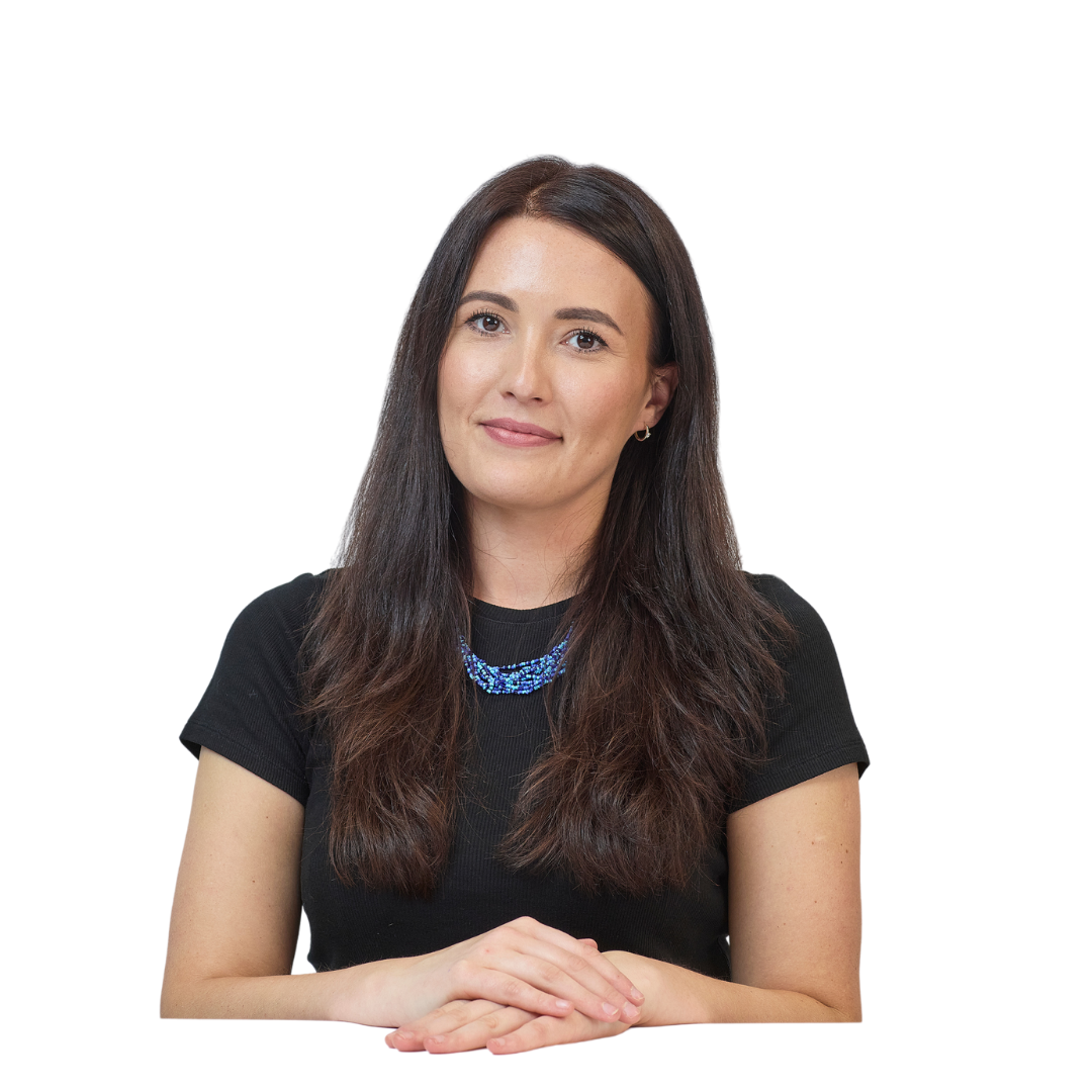 A woman with long brown hair and a blue necklace, wearing a black shirt, sits with hands folded against a plain white background.